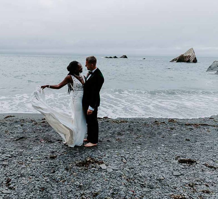 Bride in v neck sleeveless lace Pronovias wedding dress, cathedral length veil and silver sparkly bridal jewellery and groom in bottle green velvet grooms blazer standing on the shore at Tunnels Beaches Devon wedding 