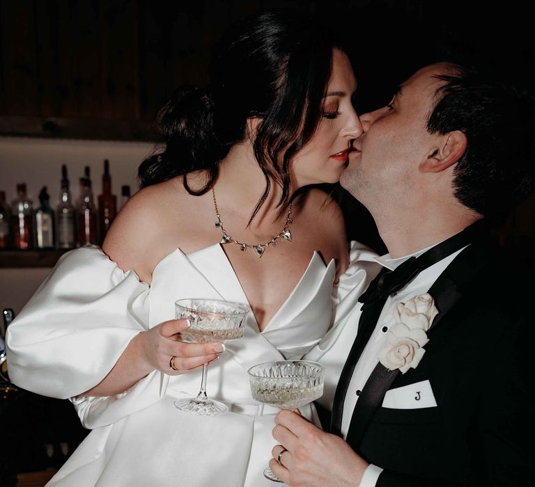 Bride and groom share a kiss whilst holding champagne coupes from the champagne tower