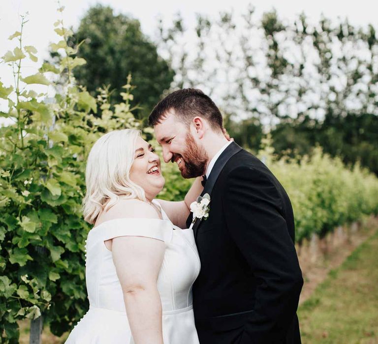 Bride in satin off the shoulder wedding dress holding white and blue eucalyptus, foliage, garden rose, gardenia, light blue delphinium and dried flower bridal bouquet tied with dusky blue ribbon standing in the vineyard with groom in classic black grooms three piece tuxedo with white rose boutonniere and black bowtie 