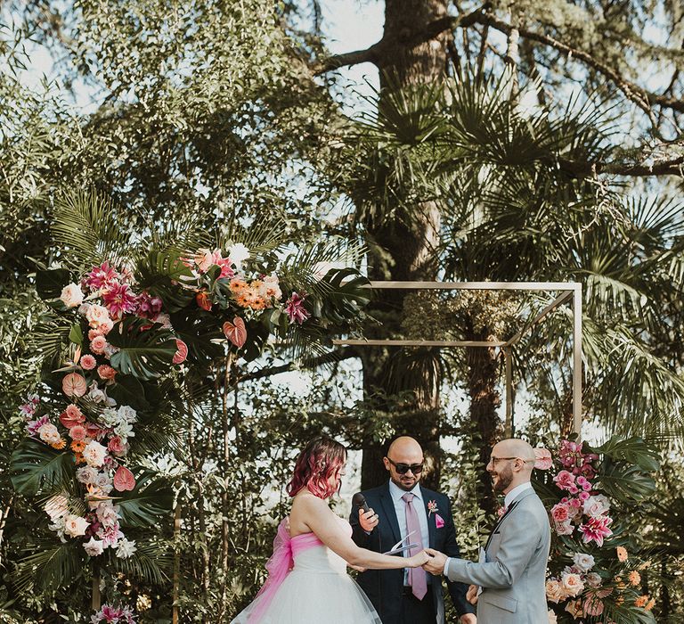 bride in a layered wedding dress and groom in a grey suit exchanging vows at outdoor Italian villa wedding venue with tropical flower arch