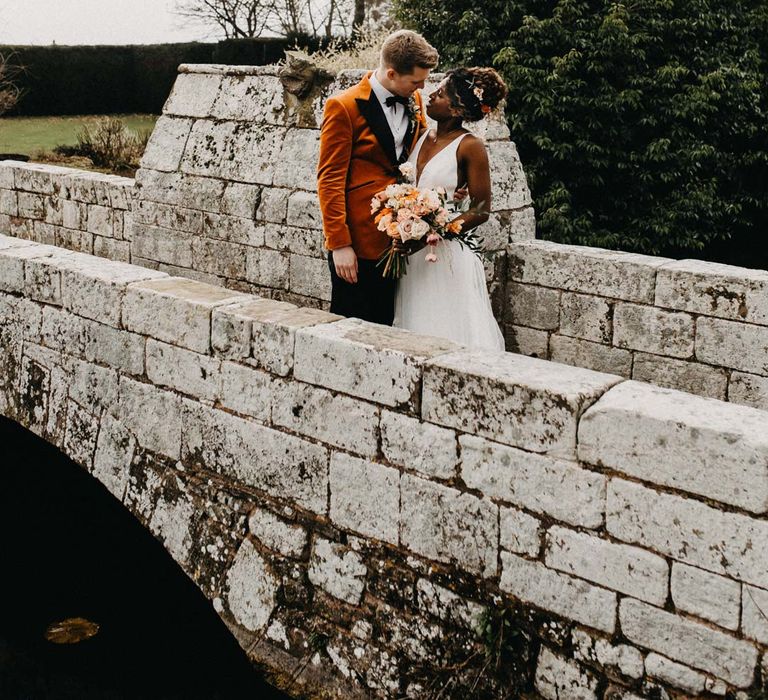 Groom in crushed burnt orange velvet grooms blazer, black bowtie and black suit trousers embracing bride in v-neck sleeveless wedding dress with 3D floral applique train holding bridal bouquet on bridge at Brinsop Court wedding venue