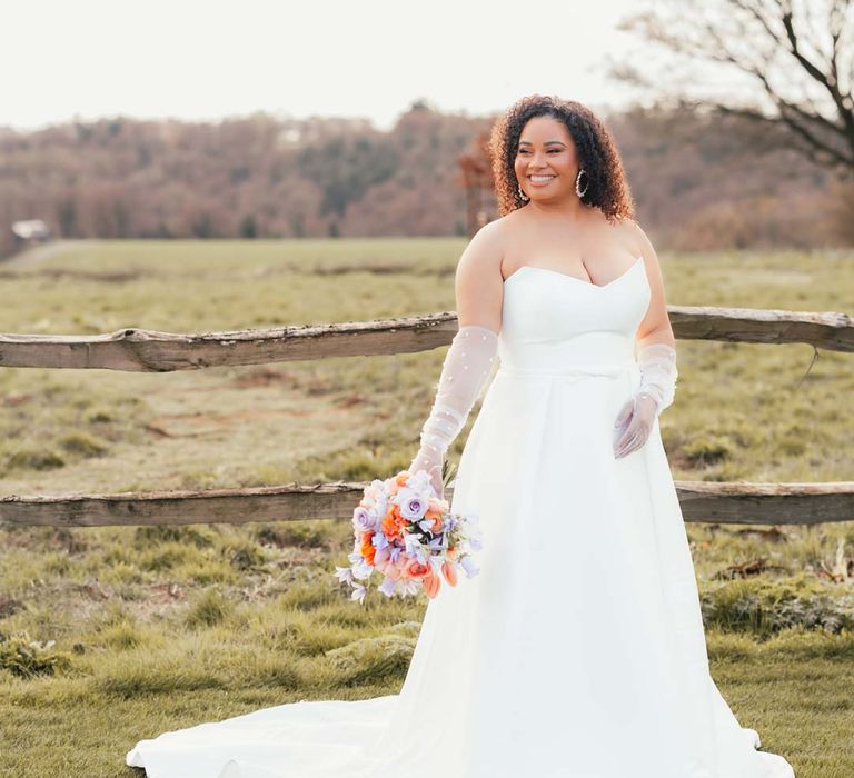 Bride in strapless wedding dress with front slit, puddle train and mesh pearl gloves holding orange, lilac and peach mixed flower bridal bouquet at Rackleys Barn wedding venue