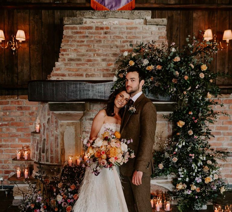 Bride in strapless ivory fringed crepe gown and chapel length veil holding large mixed bridal bouquet with garden roses, carnations, peonies and foliage standing with groom in chocolate brown grooms suit 