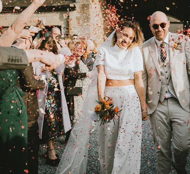 Groom in three piece cream wedding suit with sunglasses walking with the bride between their guests as they throw confetti 