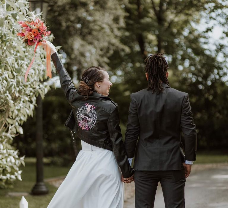 Bride in strapless sweetheart neckline wedding dress and personalised bridal leather jacket holding hands with groom in classic black tuxedo 