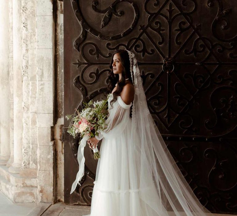 Bride in off the shoulder layered tulle wedding dress and floral embroidered cathedral-length veil holding large mixed flower bouquet 