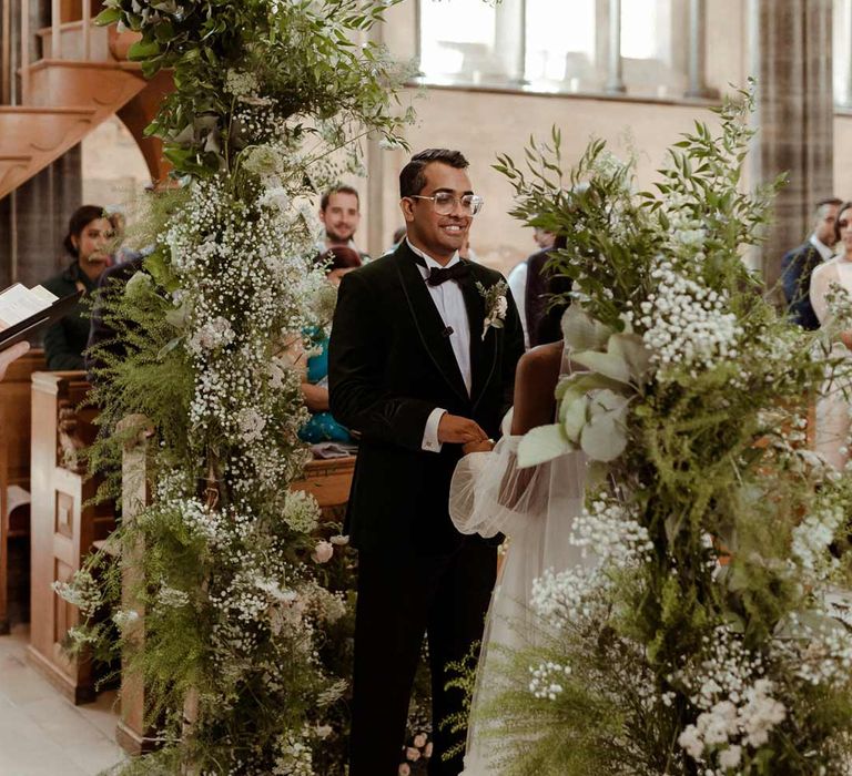 Groom in bottle green velvet grooms suit standing in Inner Temple Hall by large foliage and white flower wedding arch