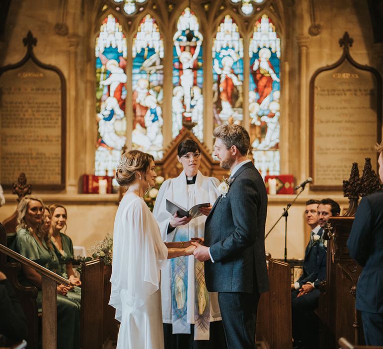 The bride and groom stand facing each other for their wedding ceremony in front of stained glass windows 