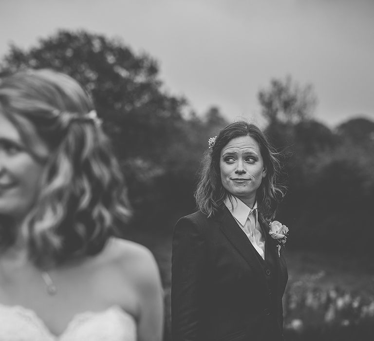 bride with bobbed brown hair wearing a suit in the background with her bride in the foreground in a strapless lace wedding dress