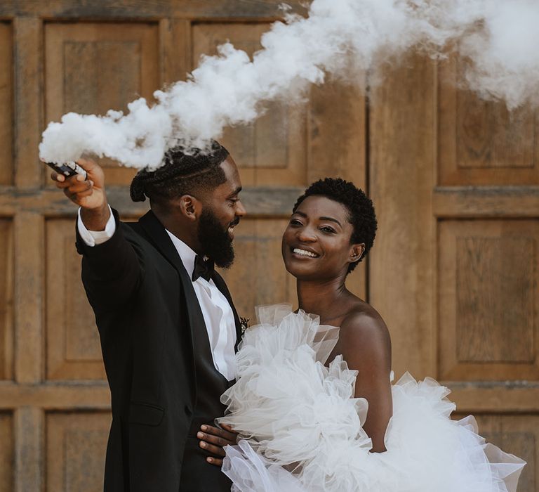 Millia London wedding dress on bride with groom in black tuxedo waving white smoke bomb