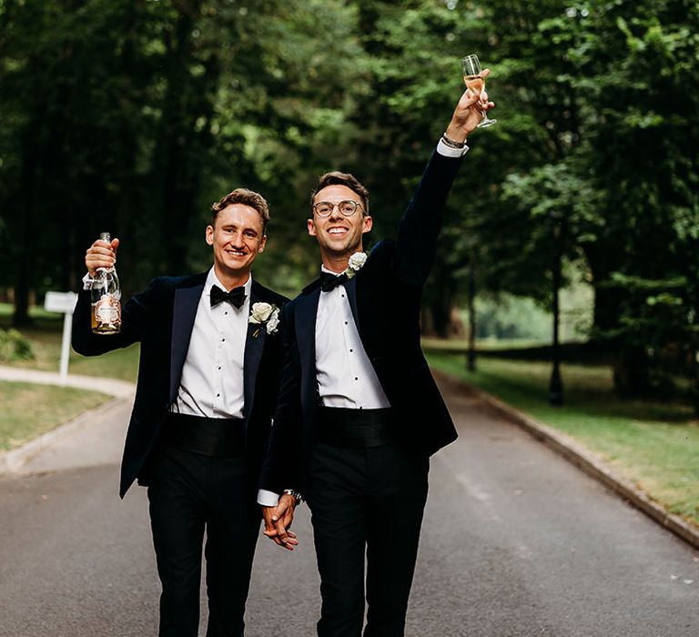 Same sex gay wedding for two grooms in navy tuxedo outfits drinking champagne 