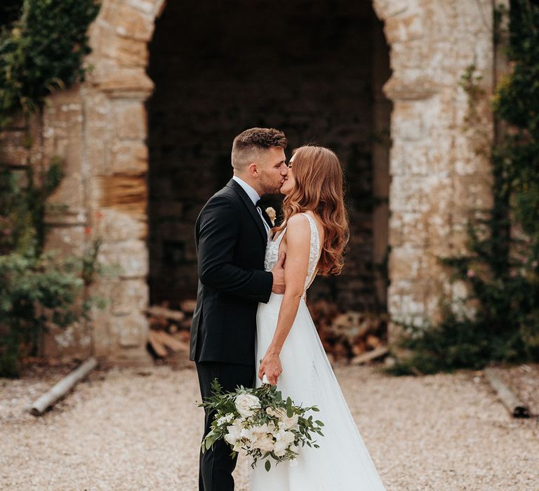 Bride carrying white rose wedding bouquet facing the groom in a black tuxedo 