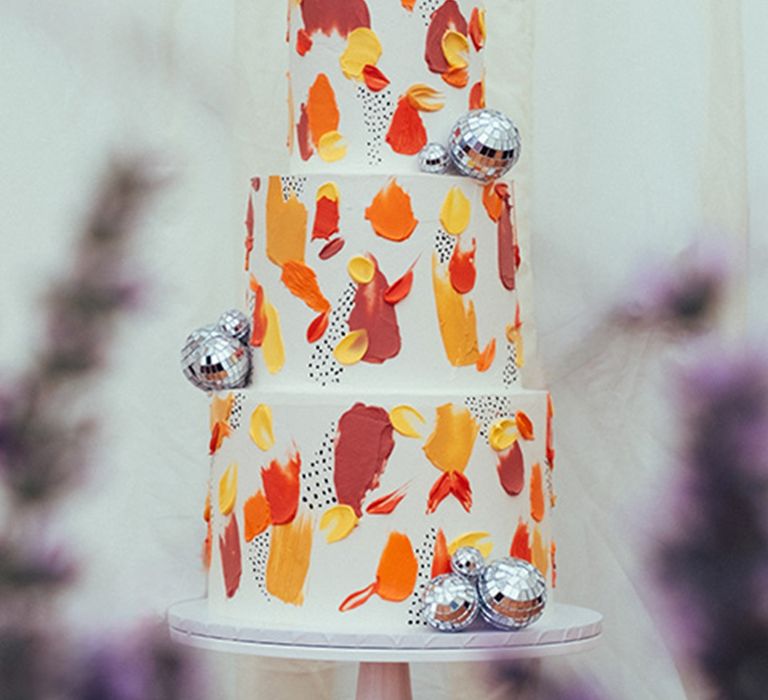 Three tier white iced wedding cake with red, orange and yellow paint style icing with disco balls 