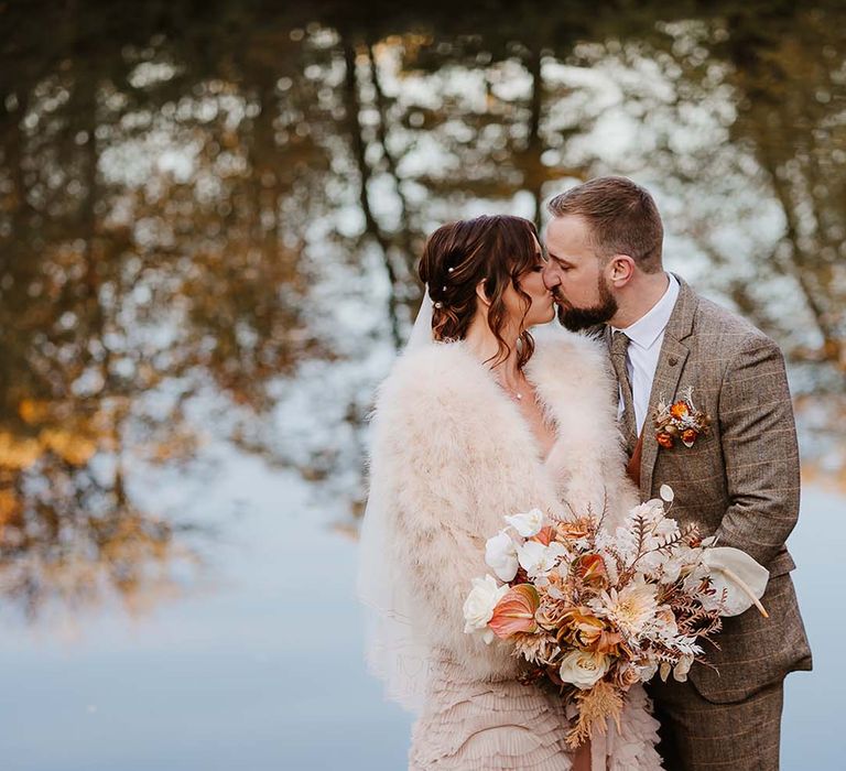 Bride in ruffle wedding dress with fur jacket for chilly autumnal wedding with the groom in brown wedding suit 