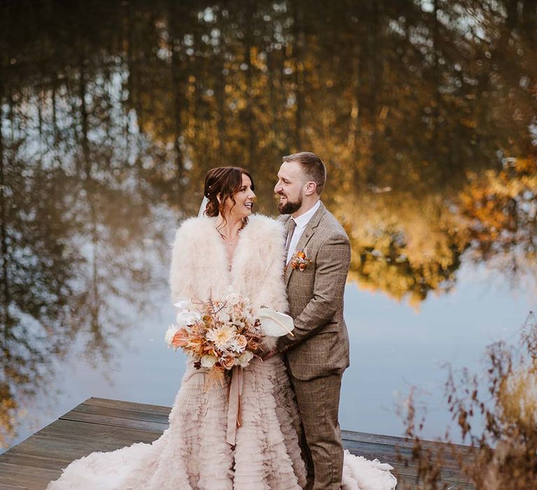 The bride wears a cream fur coat over her ruffle wedding dress from Recycle My Wedding with the groom in a brown suit at their November wedding at Styal Lodge