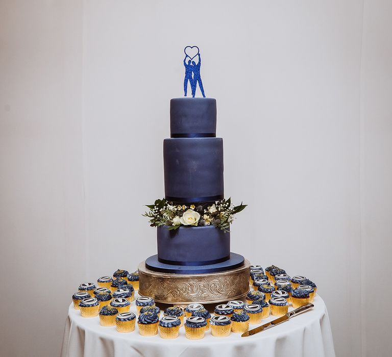 Three tier navy blue wedding cake decorated with blue glitter cake topper surrounded by cupcakes 