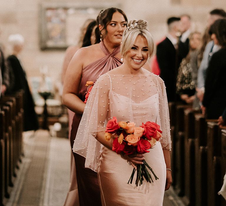 Bridesmaids in pink bridesmaid dresses walking down the aisle with colourful bouquets and pearl accessories 