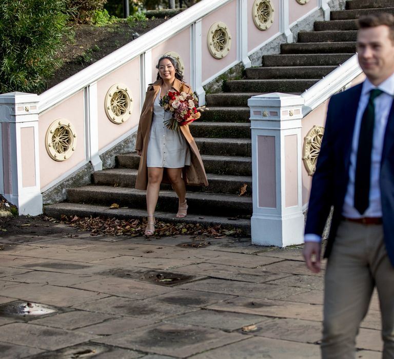 Bride in short wedding dress and beige coat walking towards her groom for their first look 