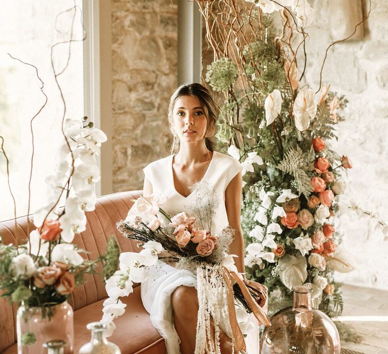 Beautiful bride sitting on a pink velvet love seat holding her dried flower and rose wedding bouquet 