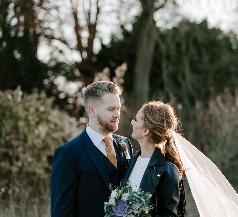 Groom in a navy suit looking at his bride in a leather jacket and cathedral length veil from Britten Weddings 