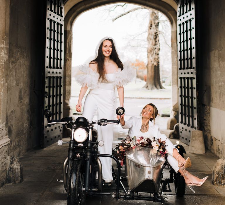 Bride in a strapless jumpsuit with ruffle shoulders and cathedral length veil standing on a motorbike with her bride sat beside her in a sidecar