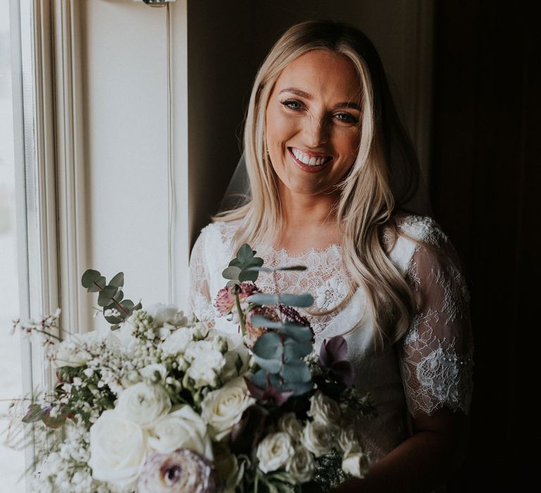 Bride in white Charlie Brear wedding dress and white Augusta Jones lace top stands by window holding white rose, ranunculus and fern wedding bouquet 