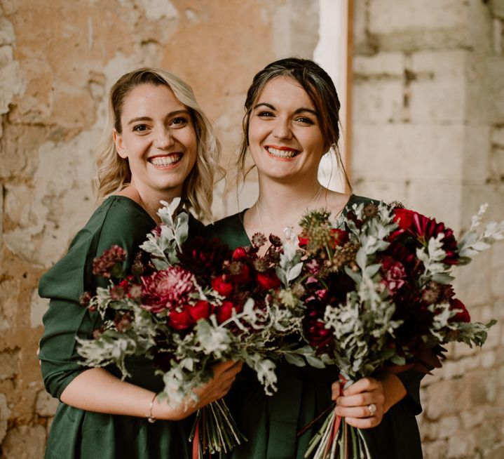 Bridesmaids in dark green dresses holding red flower and foliage bouquets 