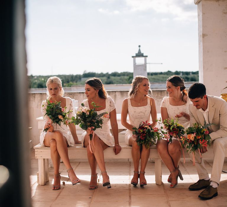 Mixed gender bridal party wearing white outfits and carrying bouquets of orange flowers and olive leaves