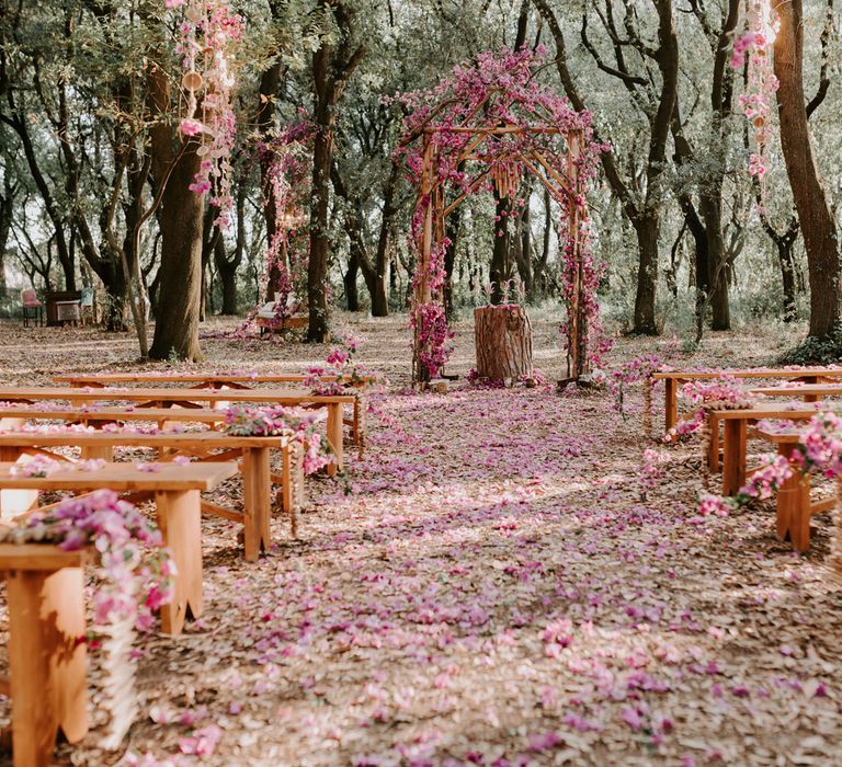 Wedding ceremony set up in Italian forest with wooden benches, wooden arch and tree stump altar all decorated with pink flowers and petals