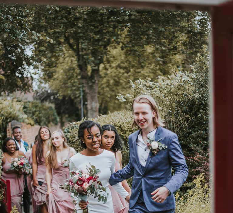 Groom in a blue suit and bride in a fitted minimalist wedding dress walking to their marquee reception followed by their bridesmaids in pink dresses