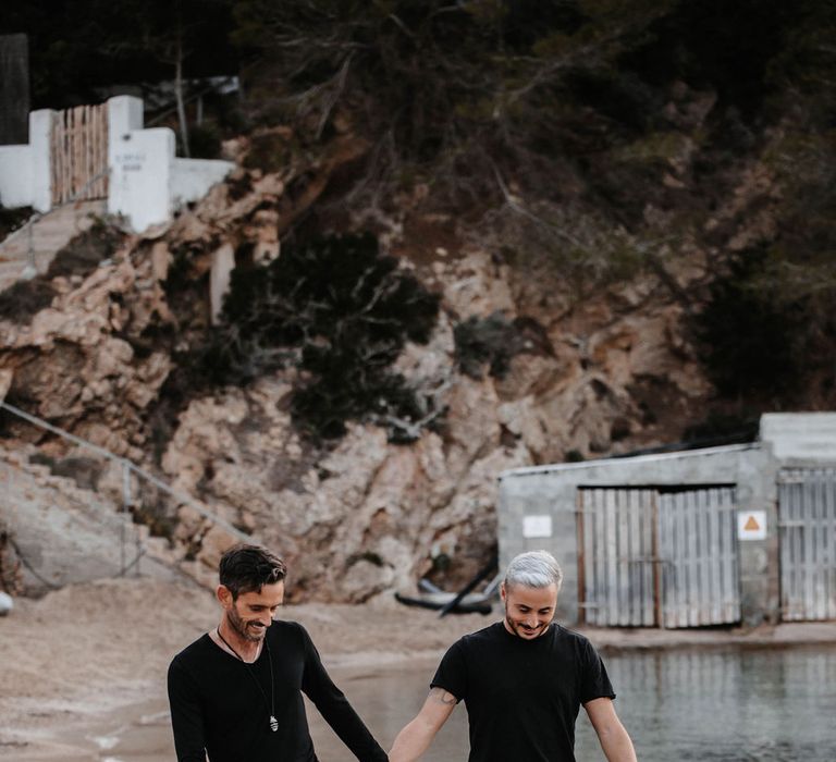 Grooms walk side by side as they hold hands and stroll along the beach