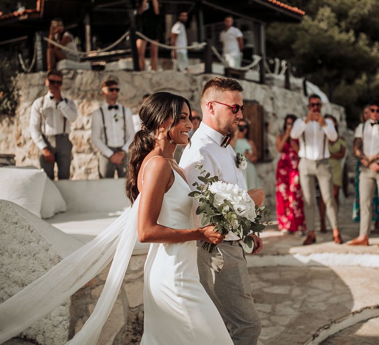 Bride in Wattea train dress and ponytail walks down the aisle clutching white flower bouquet