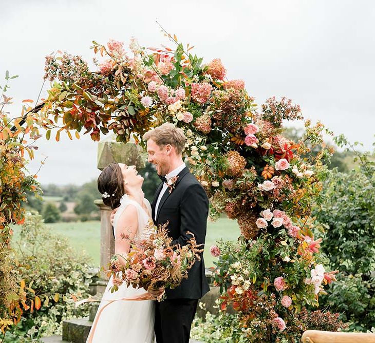 Bride and groom laughing in front of their autumn flower arch at Dorfold Hall