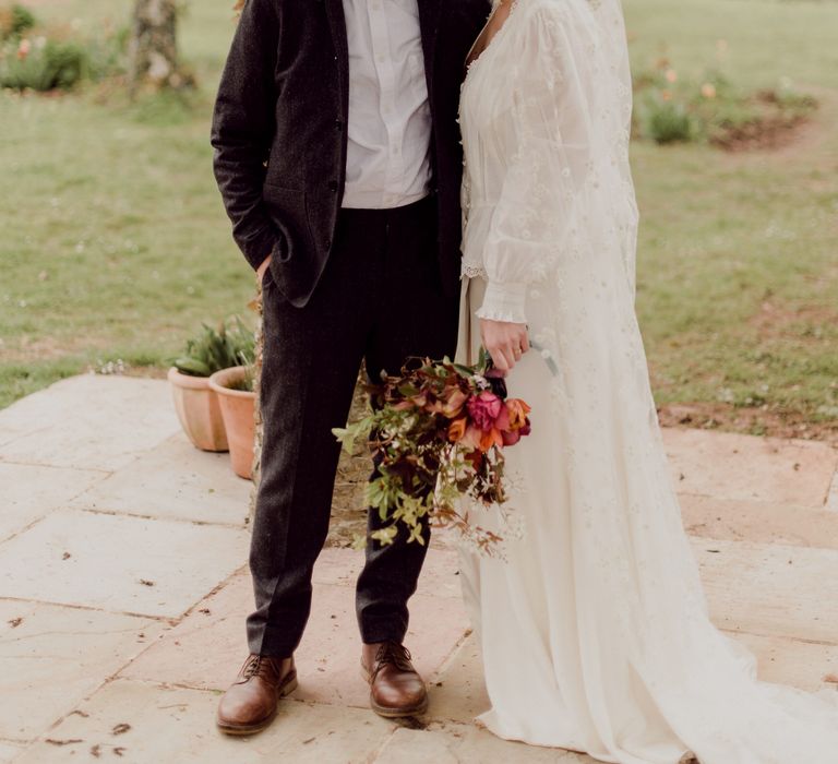 Bride in white Charlie Brear wedding dress with slit and chapel length daisy applique veil holding multicoloured wedding bouquet stands arm-in-arm with groom in grey tweed suit at garden party wedding in Devon