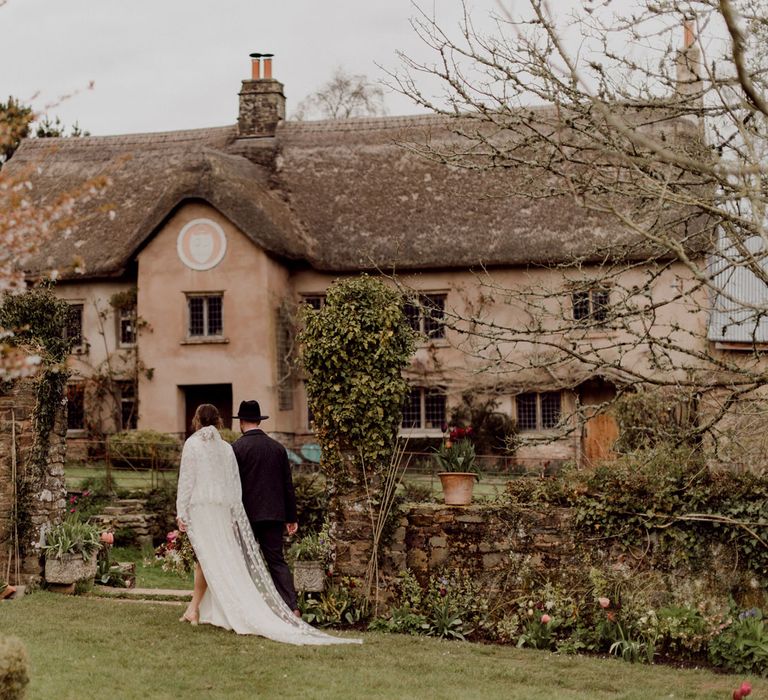 Bride in white Charlie Brear wedding dress with slit and chapel length daisy applique veil walks arm-in-arm with groom in grey tweed suit towards garden steps at garden party wedding in Devon