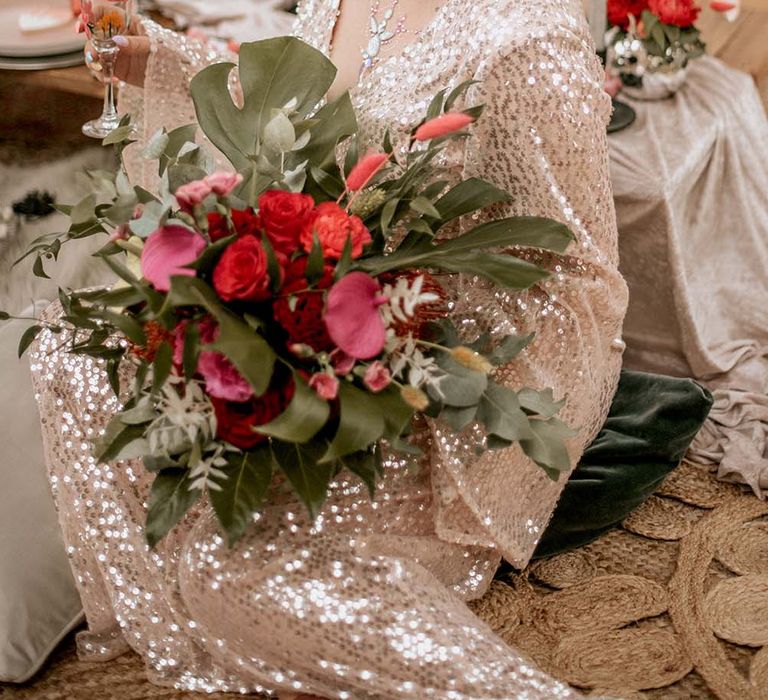 Bride in long sleeved sequin wedding dress wearing white and silver celestial headband sits holding bouquet and champagne flute at festival themed wedding