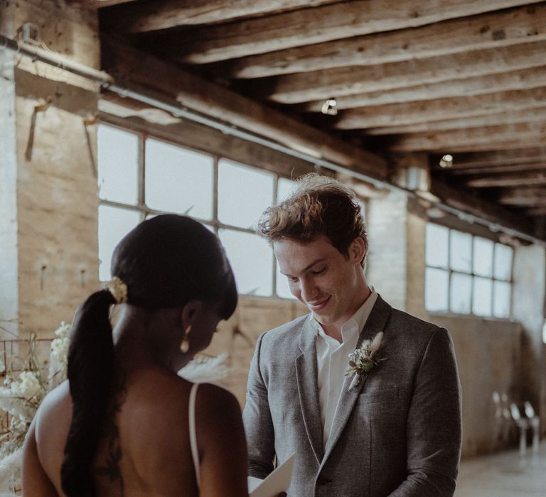 A. bride reads her vow to a groom. He wears a grey suit and looks down coyly.