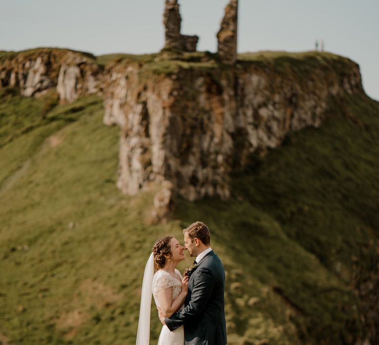 Bride in lace top wedding dress with satin skirt and veil embraces groom on clifftop after Dunluce Castle wedding