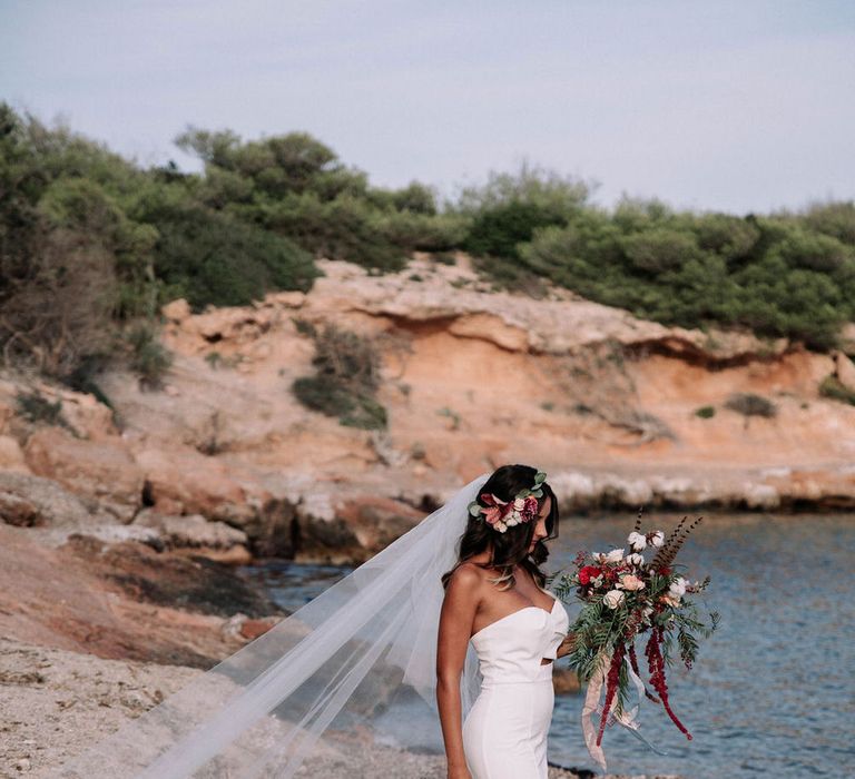 Bride walking along the beach in a strapless fitted Jarlo London wedding dress, with long flowing veil and a red bouquet