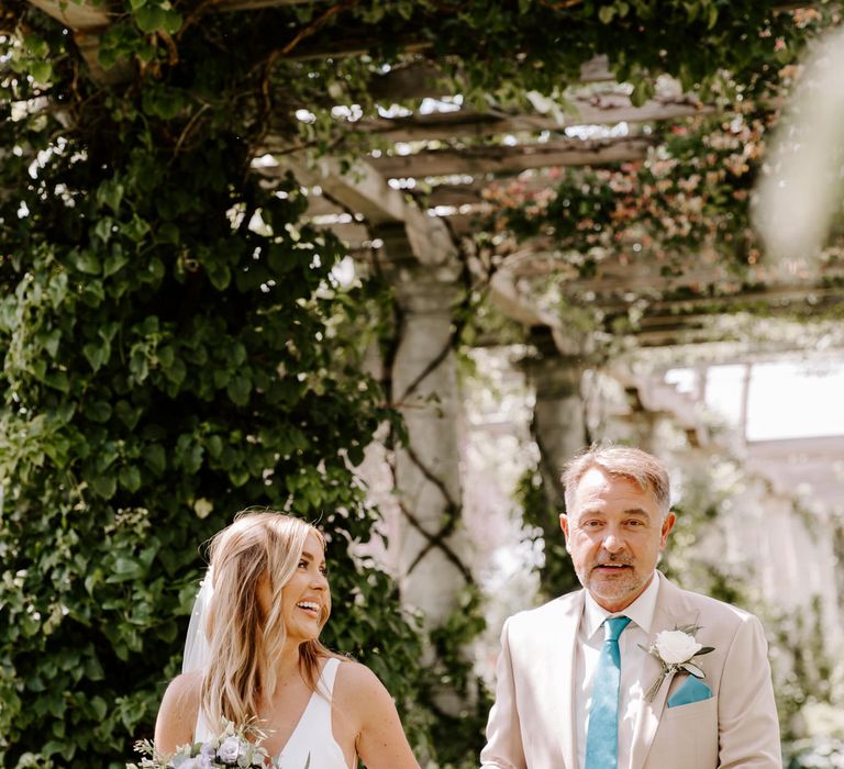 Bride walks with her father down beneath the pergola with hanging wisteria as she carries lilac bouquet