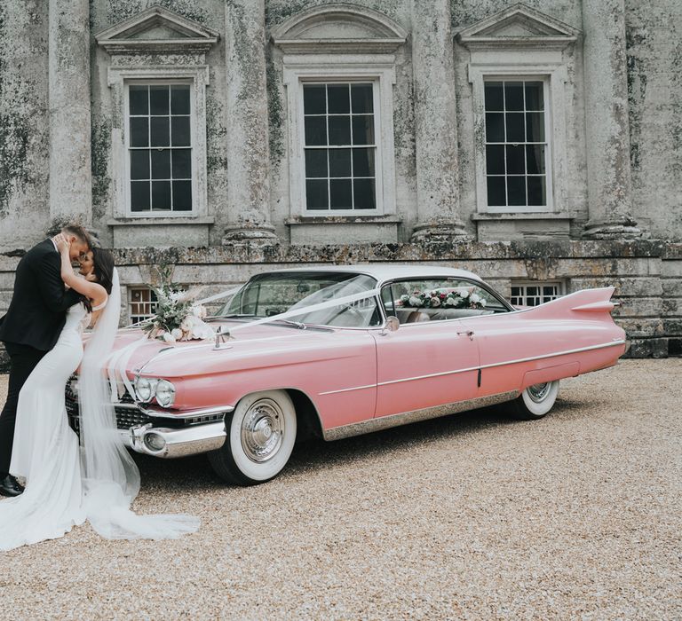 Bride in white Pronovias wedding dress and veil wraps arms around groom in black suit whilst leaning back on pink Cadillac in front of Came House Dorset after their wedding