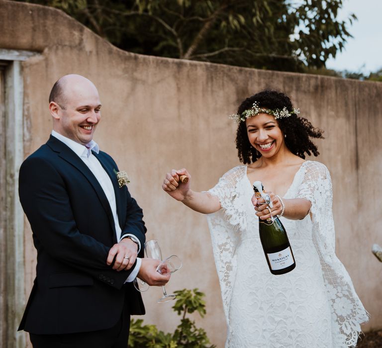 Bride pops cork of champagne bottle on her wedding day as groom stands beside her outdoors as they both laugh together