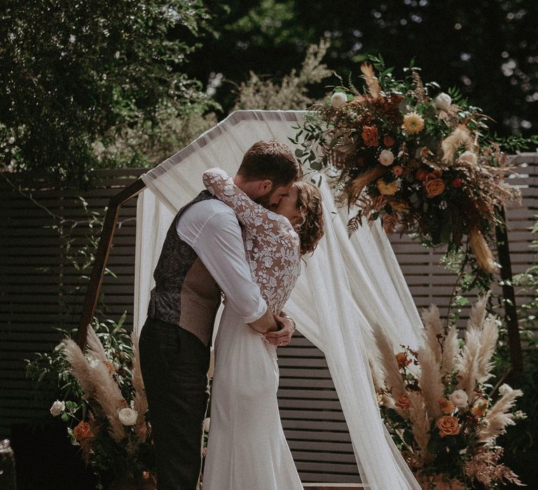 Bride in white Rime Arodaky dress, white mesh embroidered top and veil embraces groom in light brown waistcoat and white shirt with rolled sleeves in front of boho wooden wedding arch at garden ceremony with burnt orange wedding theme