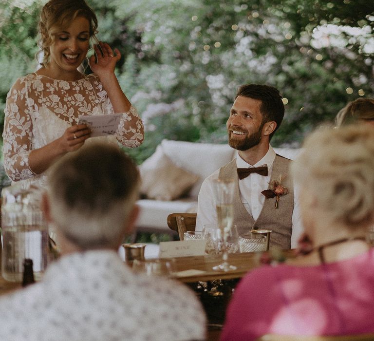 Bride in white Rime Arodaky wedding dress with mesh floral embroidered top makes speech at wedding breakfast whilst groom in waistcoat smiles up at her during garden wedding reception with burnt orange wedding theme