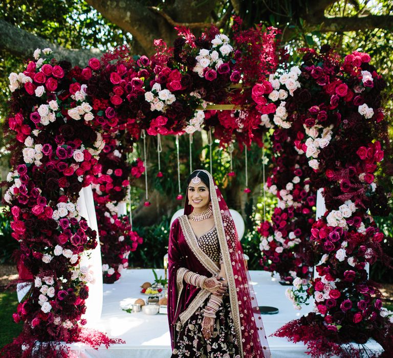 Bride stands under bright red and pink floral archway wearing traditional lehenga 