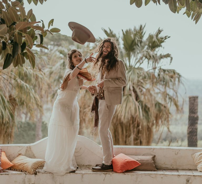 Bride throws grooms hat after wedding ceremony as bride wears boho chic gown and groom laughs 