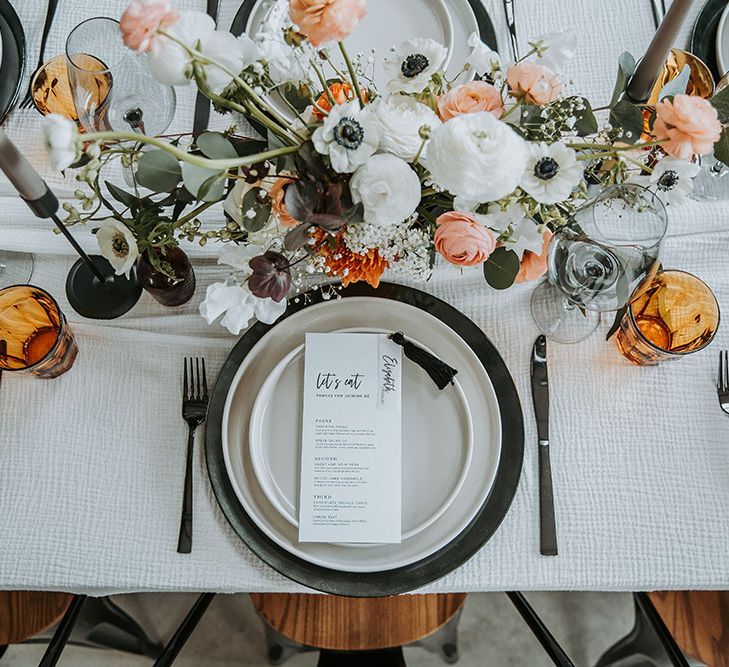 Place setting with black and white tableware and menu card, and orange, white and green flower centrepiece decor