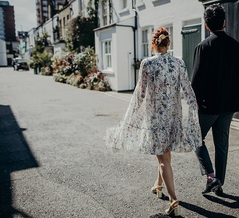 East Asian bride in a short wedding dress with blue floral design and long cape sleeves holding hands with her groom in a navy velvet jacket in London