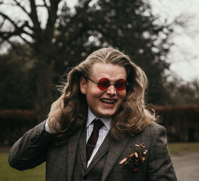 Groom in a dark grey tweed suit with long hair and rose tinted sunglasses for pub wedding