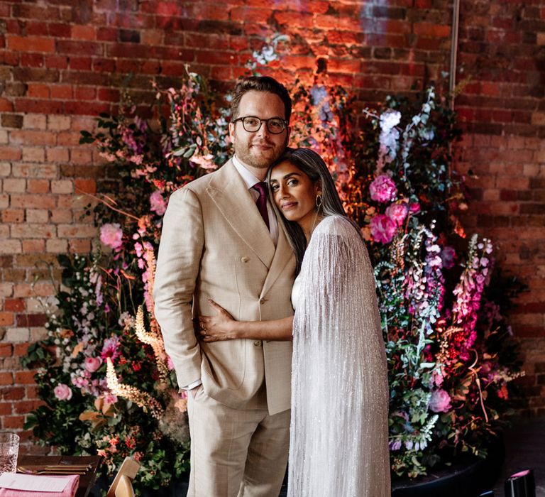 Bride in Halfpenny London tasselled bridal cape hugs groom in double breasted linen suit and gold Nike Air Force 1 trainers as they stand in front of floral installation before wedding breakfast at Loft Studios London